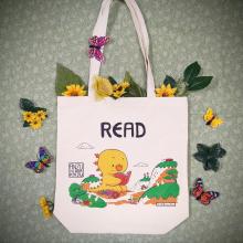 Art of Anzu a yellow kaiju reading a book in nature under the word READ on a natural canvas tote bag. The bag is placed on green fabric with fake flowers and butterflies around it.