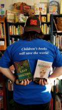 Apollo holding one book in each hand behind their back under the logo on their blue shirt "Children's books will save the world!"