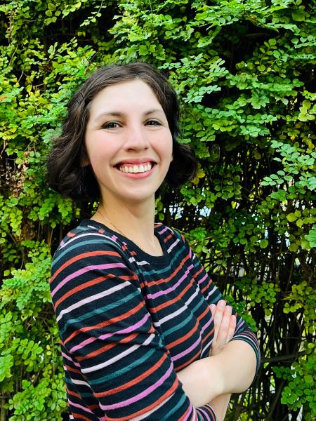 Emma Steinkellner author photo. A white woman with short brown curly hair cut in a bob wearing a pink, white, red, and green striped shirt standing with her arms crossed in front of greenery.