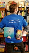 Iz holding one book in each hand behind their back under the logo on their blue shirt "Children's books will save the world!"