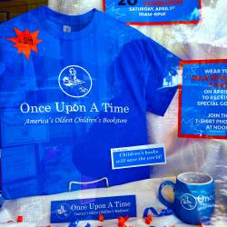 The shirt on display against a white background next to signage. A blue bookmark and Once Upon A Time mug sit in front of the shirt. 