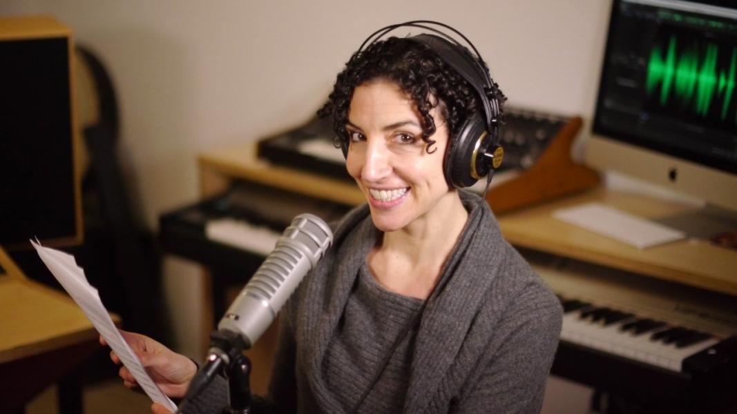 Rebecca Sheir author photo. A white woman with short, brown curly hair wearing headphones in front of a microphone wearing a grey sweater. 