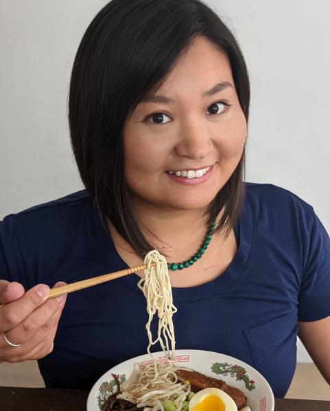 Shiho Pate illustrator photo. An Asian woman with short strait black hair in a bob cut smiling as she eats from a bowl of ramen. She is wearing a navy blue shirt with a green beaded necklace.