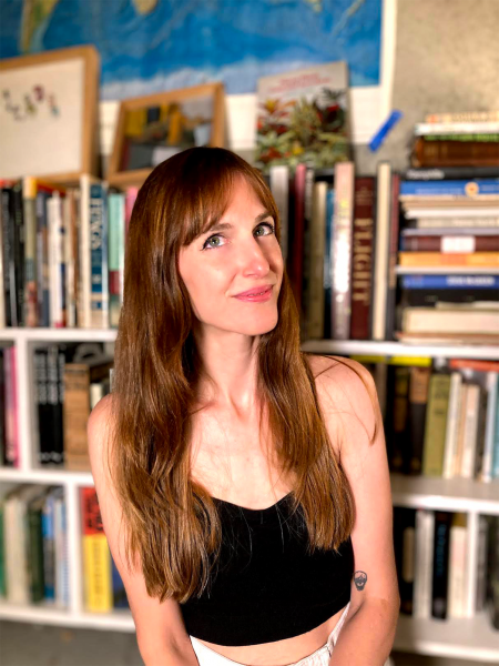 Leah Hayes photo. A white woman with long straight brown hair stands in front of a bookshelf in a black tank top and white pants. 